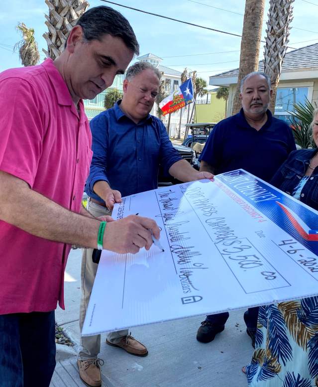 A man in a pink shirt signs a giant check while three others hold it up.