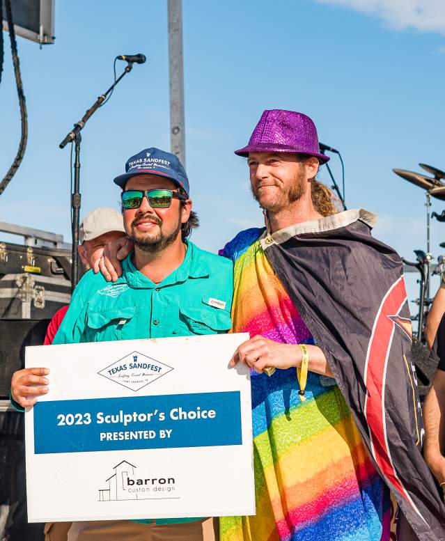 Two men, one draped in multicolored flags and a purple fedora and the other in a teal fishing shirt and hat, pose with a sign that reads "2023 Sculptor's Choice"