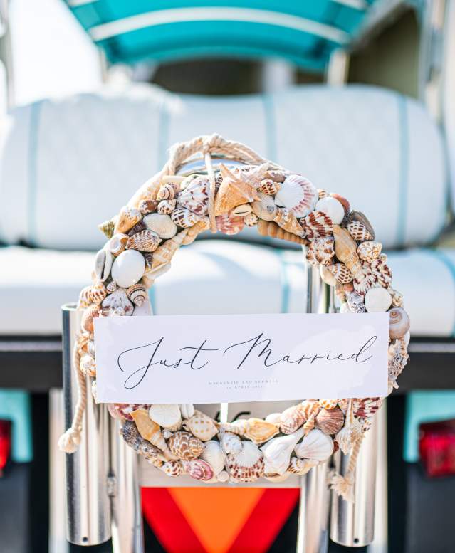 In focus is a wreath made of seashells with a placard reading "Just Married" in script font. The wreath is attached to the back of an out-of-focus turquoise golf cart