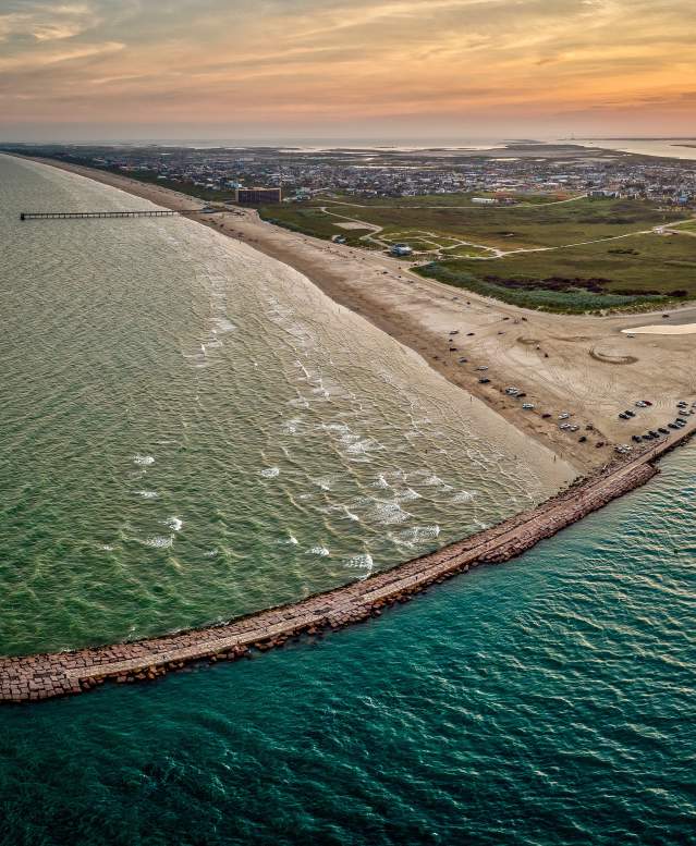 An aerial view of the coast showing the jetties in a teal water with the sun setting in the distance