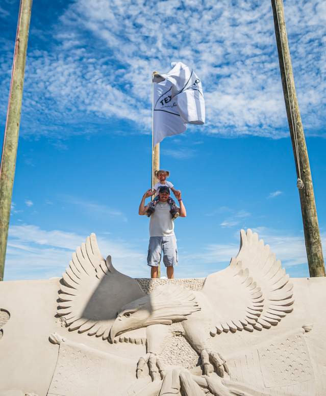 A man carries his toddler daughter on his shoulders and they pose atop a large sand mountain carved with an eagle