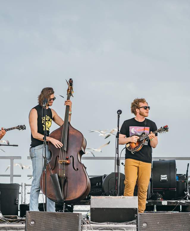 A band of four people—one with a banjo, cello, ukelele, and guitar—stands on stage on the beach.