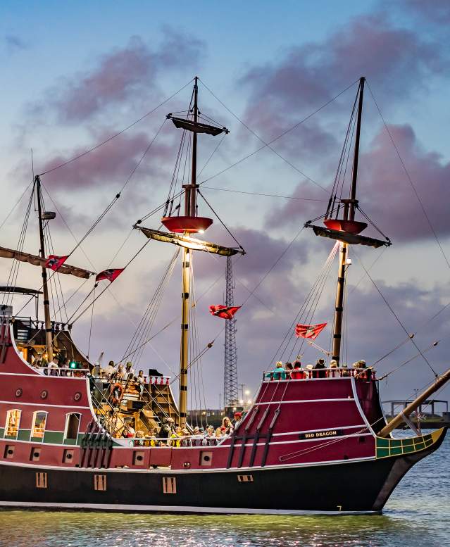Large red pirate ship in the ocean with lights on in facing the deck. The sun has almost set and gives the sky and clouds a purple tint.