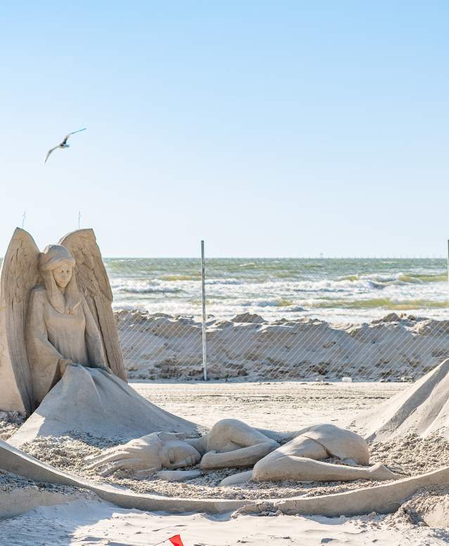 Sand sculpture depicting four angels at four corners holding a blanket. In the middle of the blanket, a woman lies curled up.