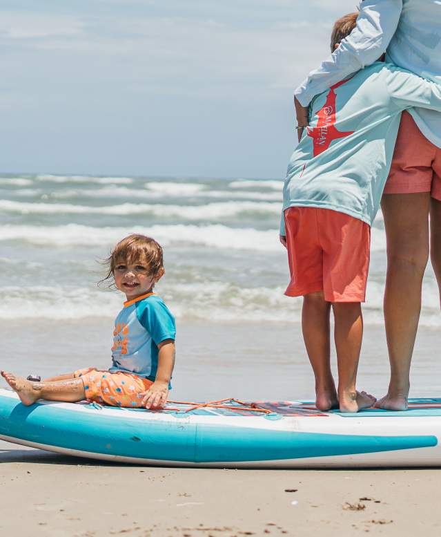 Family photos, child sitting on the end of a paddle board on the sand as family stands on top.