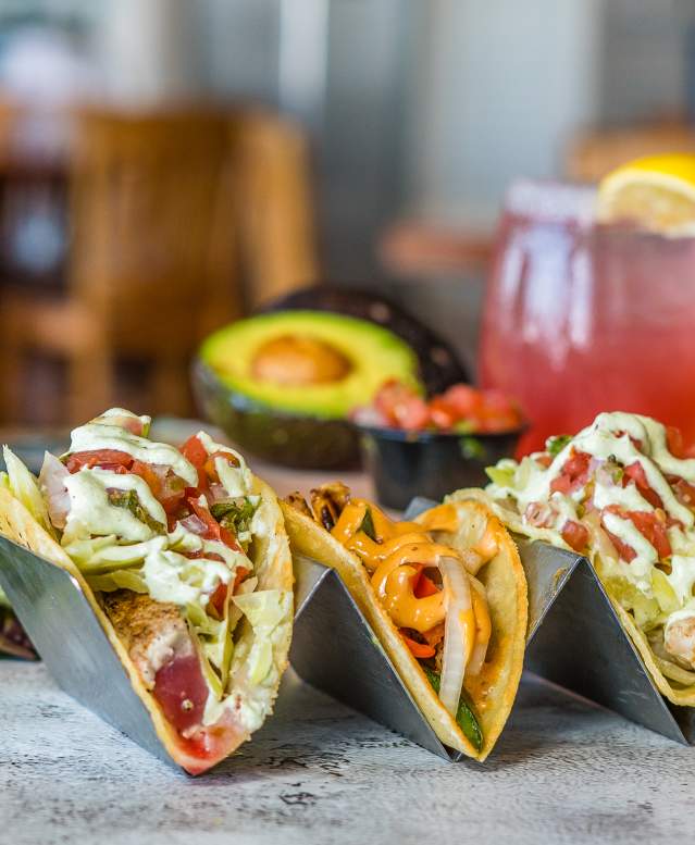 A trio of tacos sits on a table surrounded by an avocado, a pink margarita, and other ingredients