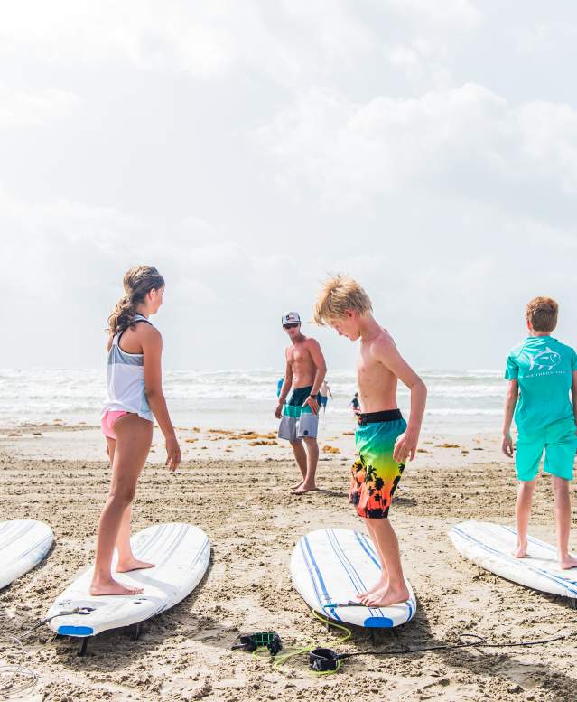 A group of five kids stand on surf boards resting on the sand while an instructor stands in front of them