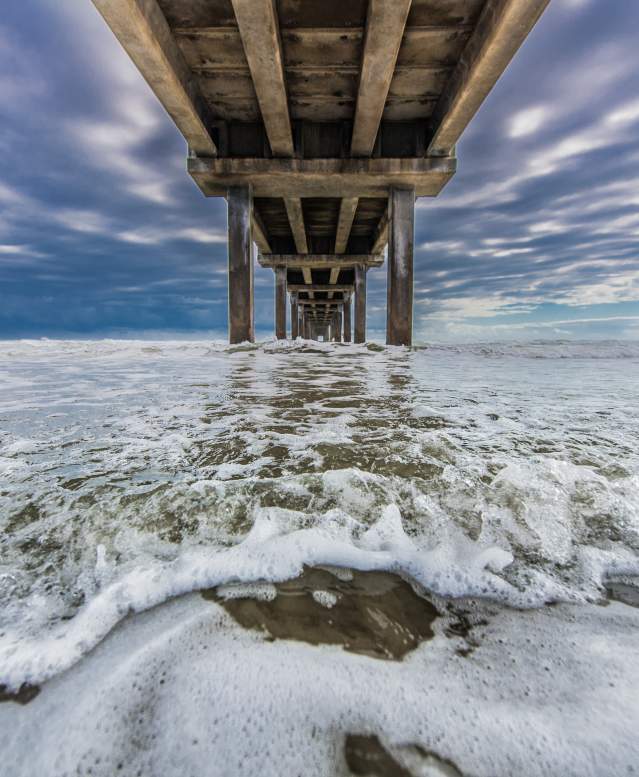 Foamy seawater rushes underneath a pier with a bright, cloudy sky