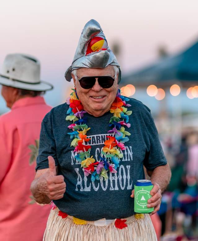 A man gives a thumbs up to the camera. He's wearing a metallic shark hat, a hula skirt, and several leis.