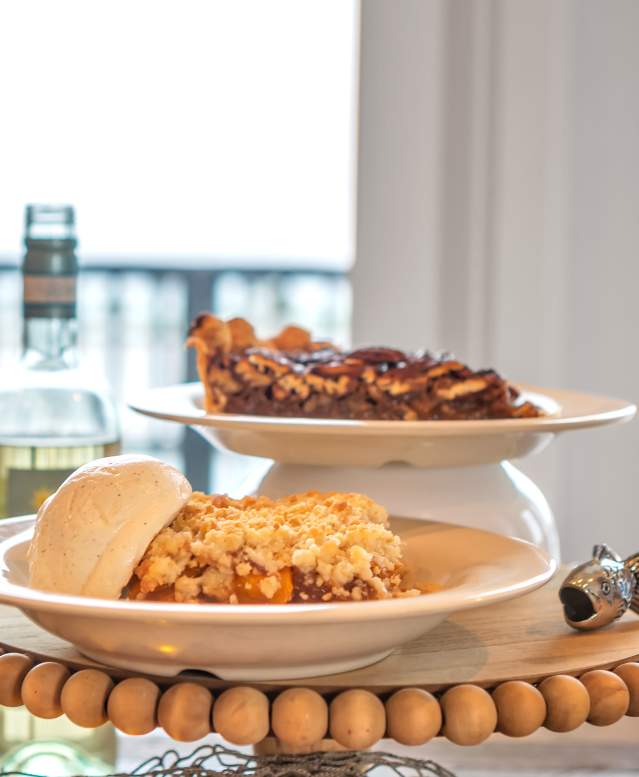 A plate of cobbler with ice cream and a plate of pecan pie sit stacked on an elevated dessert tray. To the left are open bottles and glasses of wine.