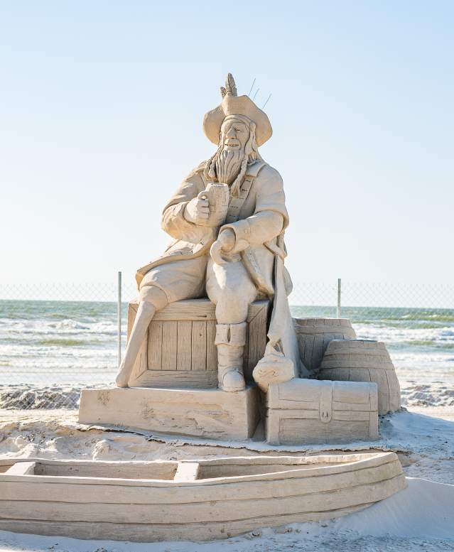 Sand sculpture of a pirate drinking out of a cup and sitting on barrels. In front of him is a rowboat made of sand