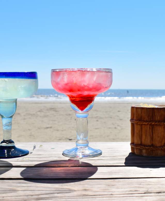 A row of margaritas sitting on a wooden table on the beach.
