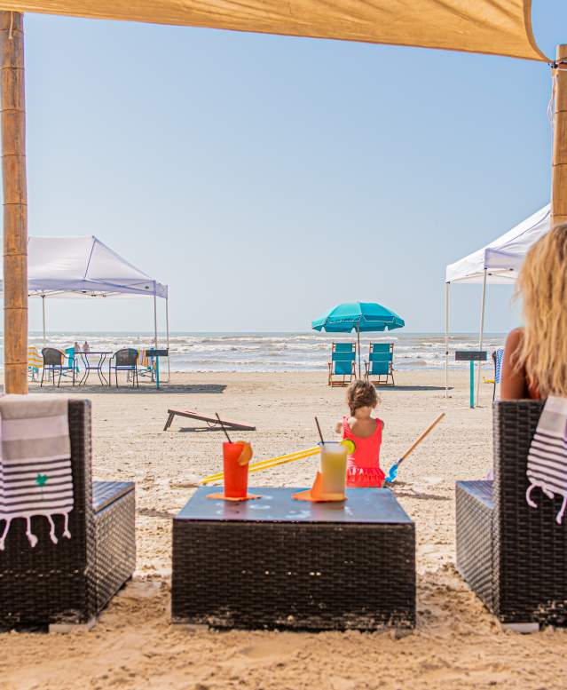 Two chairs on the beach are seen from the back. A blonde women sits in one chair and a table with tropical drinks sits between the chairs. A child plays in the sand in front of the setup.