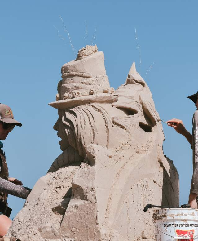 Two people, a man and a woman, stand on either side of a sand sculpture and carve details.