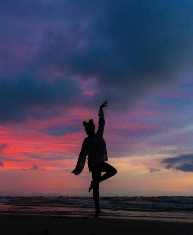 A model is silhouetted in a dancing pose against a dark pink, blue, and purple sunrise on the beach