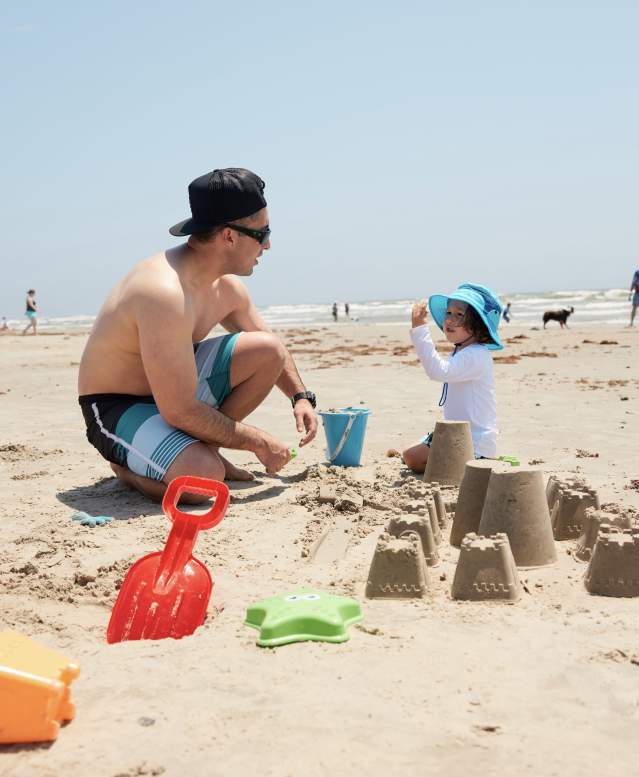 A man in a swimsuit kneels on the sand next to a child wearing sun gear and a hat. They pack sand in a blue bucket and build a sandcastle.