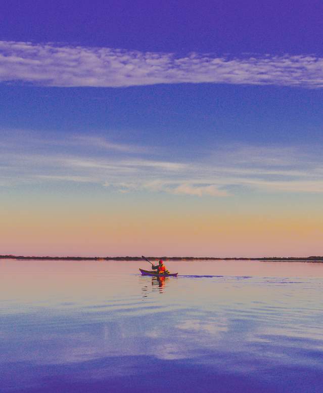 A lone kayaker wearing red floats on calm water that reflects the orange and blue sunrise sky
