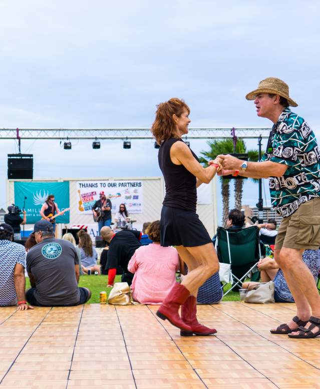 A couple dances on a dance floor in front of groups of people sitting on green grass facing a stage.