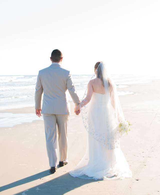 A bride and groom in wedding attire walk hand in hand, backs to the camera, down a light, bright beach