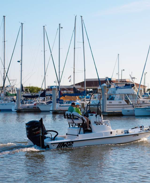 A fishing boat with two people in the driver's seat putters through a marina with rows of sailboats behind