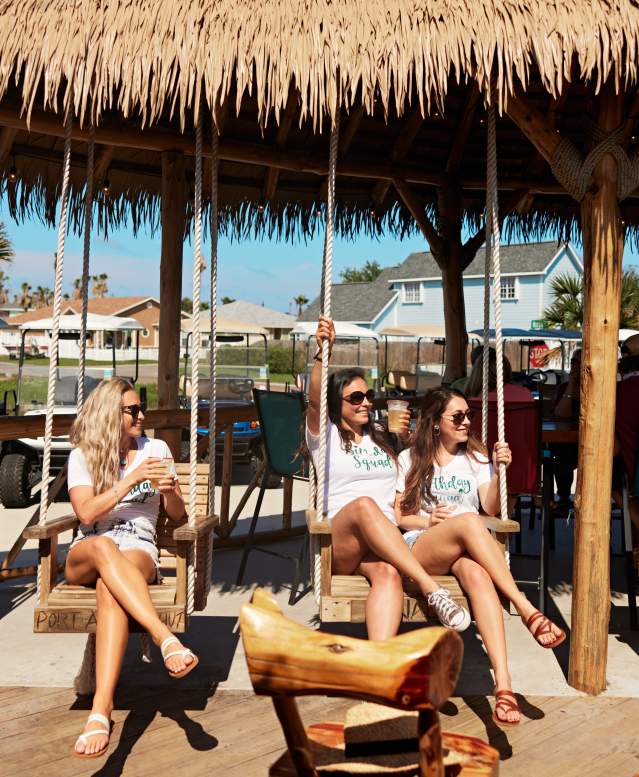 A group of five women wearing matching birthday shirts sit in tiki-style swings