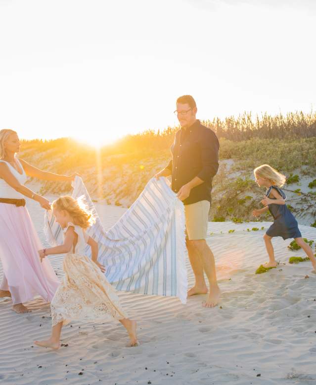 On the beach in front of sand dunes, a mother and father hold a striped blanket between them while two young girls run next to them