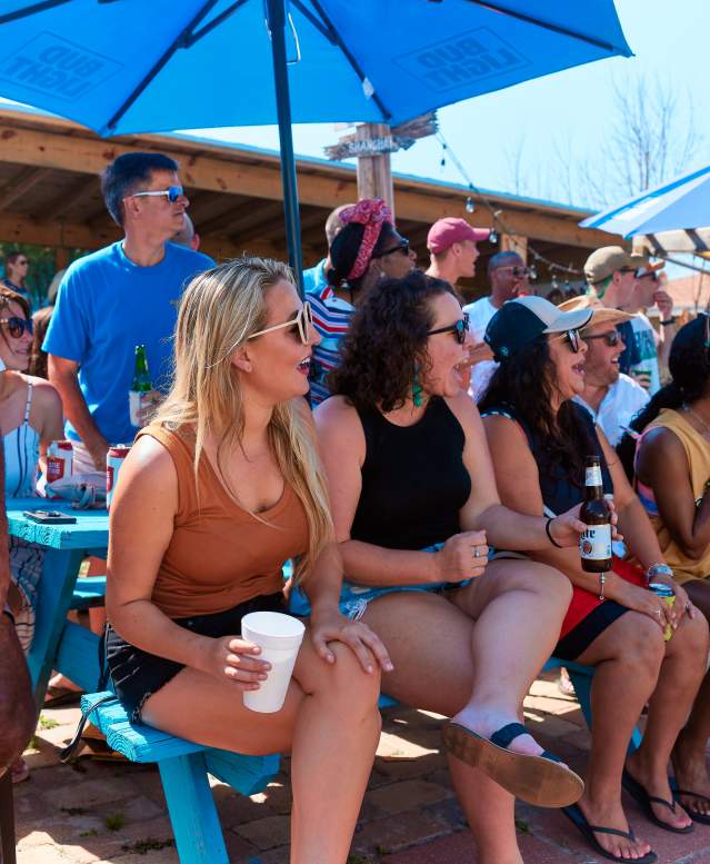 A diverse group of people sits at a picnic table with excited expressions as they watch an off-camera event