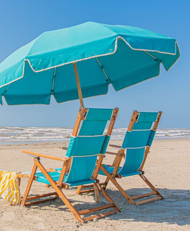 Two blue beach chairs with an unbrella between them. There is a yellow blanket over one chair and yellow beach toys near by. In the background thier are two people playing in the ocean.