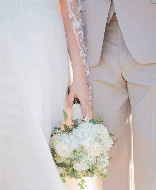 A close-up photo of a couple standing on the beach in wedding attire. The bride holds a bouquet.