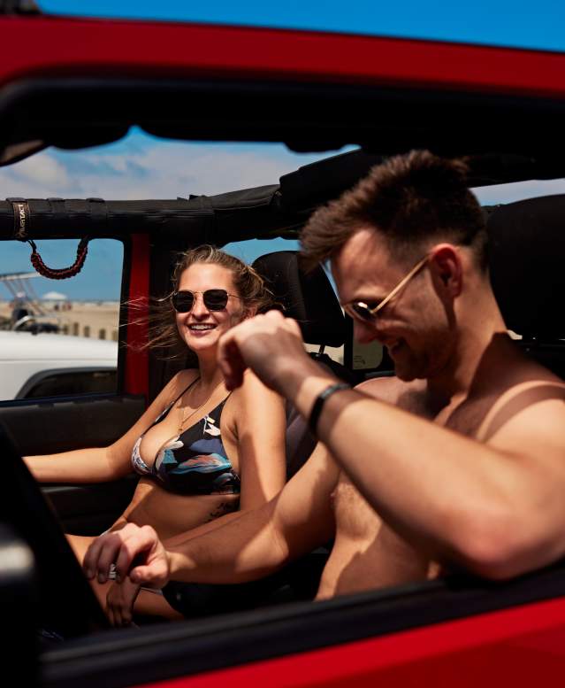 A man and woman in swimsuits are seen sitting in a red Jeep from the Jeep's window view