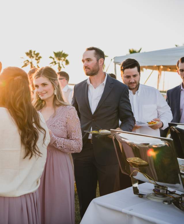 Bridesmaids in mauve dresses and men in suits wait in line at a buffet with palm trees in background