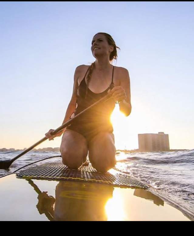 Girl kneels on a stand up paddle board in the water with a paddle in hand.