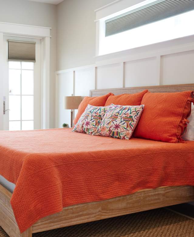 A bed with a wooden headboard and orange quilt and floral pillows sits in the middle of a bright, light bedroom.