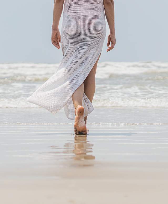 Bottom half of a woman in a long white cover up walking on the beach towards the water