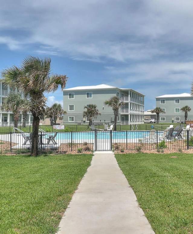 Wide shot of several sage-colored buildings set back behind a large pool with palm trees. right in front of the camera is nice green grass.