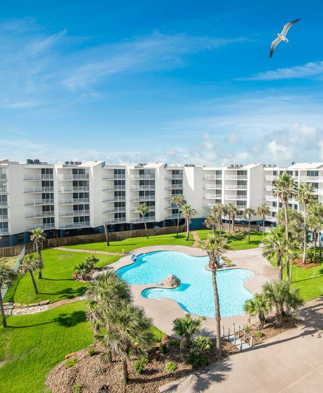 Aerial photo of a beautiful white condominium complex with palm trees, a pool, and green grass.
