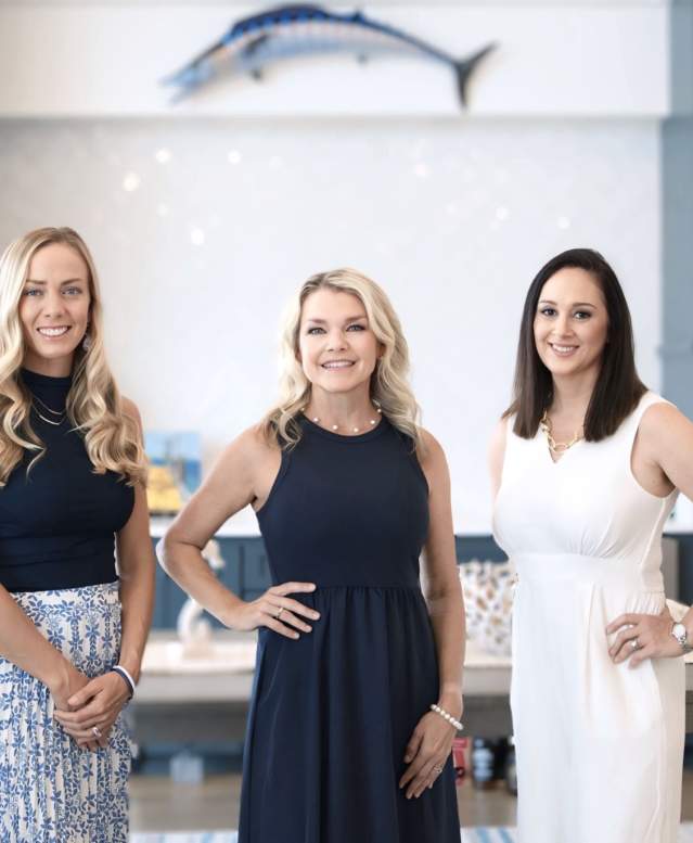 Three women in coordinating navy and white outfits stand in a coastal home and smile at the camera