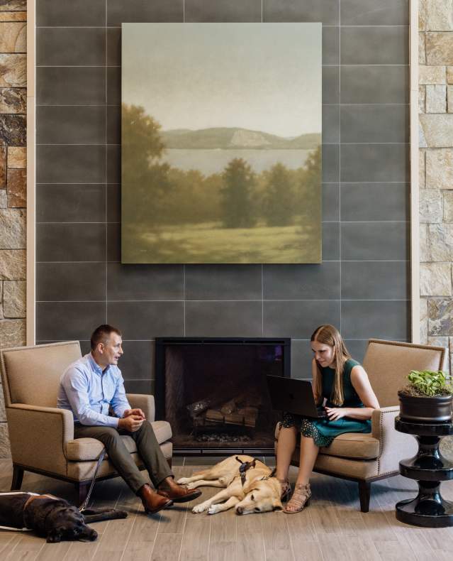 Two people are seated in front of a fireplace with their service dogs lying asleep at their feet. On person has a laptop open and is describing her report to the listener.