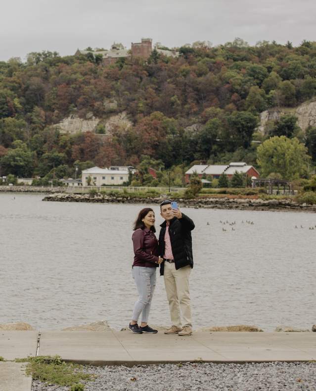 A couple stands taking a selfie in front of the Hudson River at Riverfront Green Park. Both the sky and water behind them look grey.