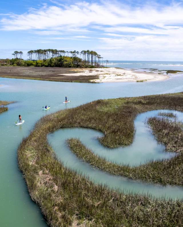 Aerial view of paddleboarders exploring winding salt marsh creeks near Waites Island in North Myrtle Beach, South Carolina, with pristine coastal scenery and clear blue skies.