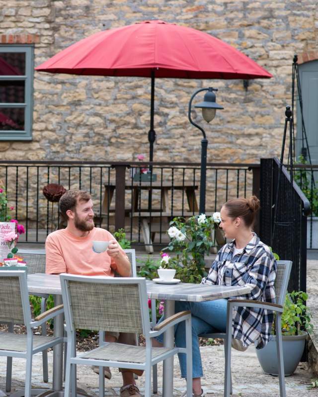 A man and a woman sat on a outdoor dining table with hot drinks in courtyard with listed barn building in background