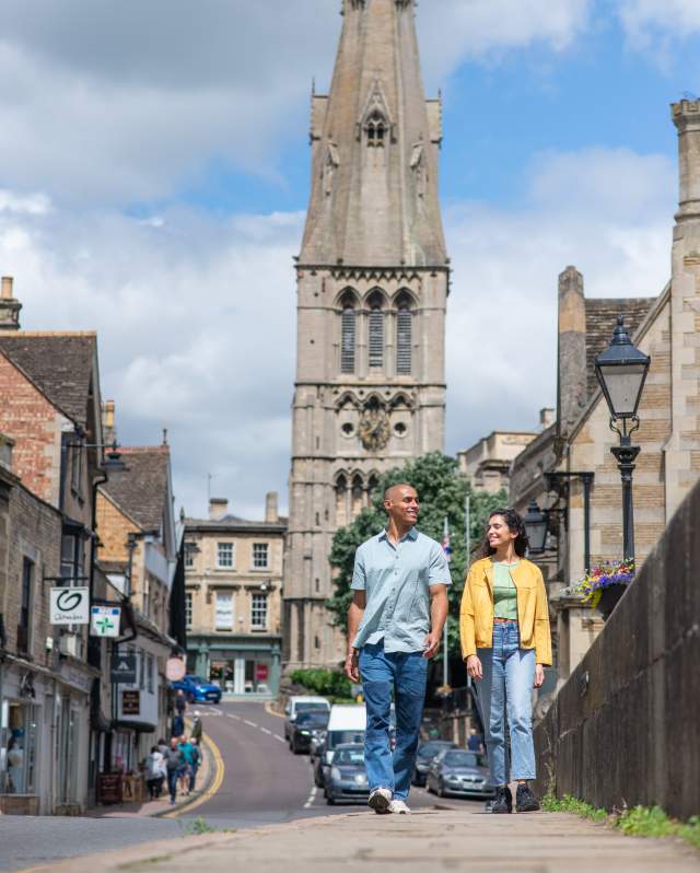 Two people walking along a pavement on a bridge in Stamford with a church in the background in sunlight