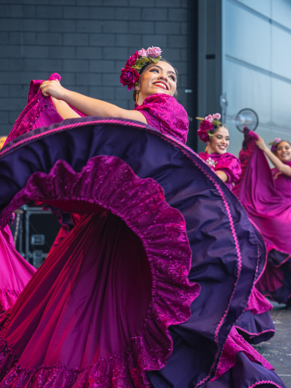 A group of female dancers performs a traditional Mexican folklórico dance on stage. They wear vibrant magenta dresses with purple ruffles and floral headpieces. The lead dancer in the foreground smiles brightly while gracefully twirling her flowing skirt. The dancers behind her mirror the movement, creating a colorful, energetic scene against a modern stage backdrop.