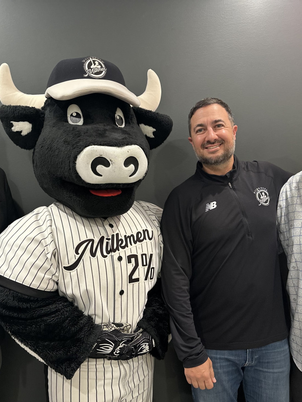 Three men pose for a photo with the Milwaukee Milkmen mascot, a smiling black and white cow wearing a pinstriped baseball uniform and cap, standing in front of a plain dark gray wall.