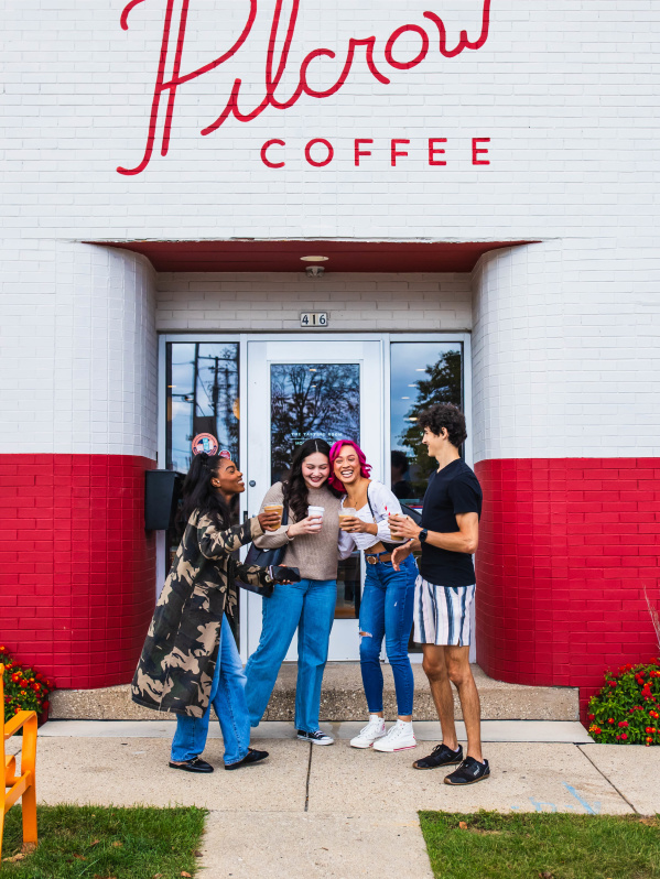Four friends holding coffee cups and laughing outside Pilcrow Coffee, standing in front of a red-and-white storefront with an “Open” sign in the window.