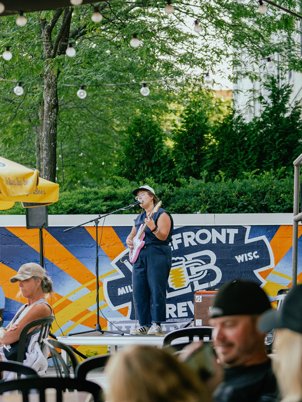 A woman in a navy blue romper singing into a microphone on a stage, holding a pink guitar, with people in front of her sitting at tables under gold and blue umbrellas on a patio along the riverfront with trees in the background.