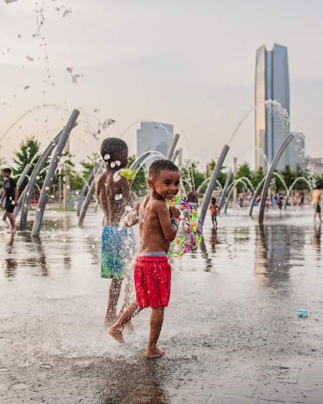 Children playing the splash pad at Scissortail Park. OKC skyline can be seen in the background.