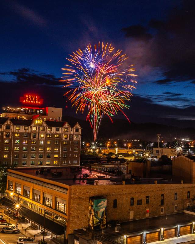 Fireworks burst in the night sky above downtown Rapid City, with the illuminated Hotel Alex Johnson and city lights glowing beneath dark clouds at dusk.