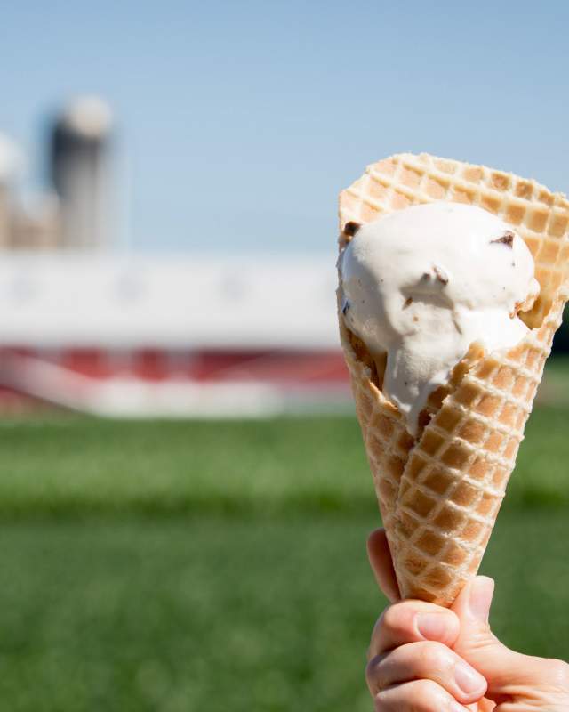 A hand holding up a cone of ice cream in front of a barn