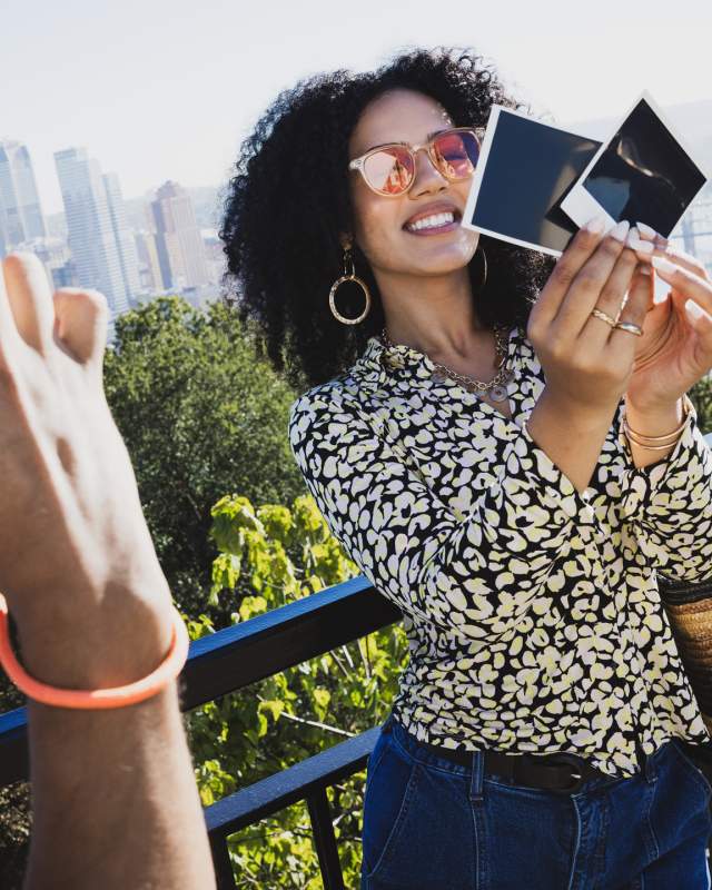 A woman holding up polaroid photos while a man takes a photo of her in front of the Pittsburgh skyline